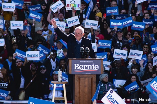 A smiling Bernie Sanders lifts up his arm in acknowledgement of the crowd